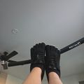 A person wearing black lace-up boots holds their feet up toward the ceiling, with a ceiling fan and light fixture visible in the background.