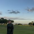 A person in dark clothing and sunglasses stands on a golf course holding a golf club, with green fairways, trees, and a partly cloudy sky in the background