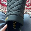 Person wearing black quilted winter gloves with yellow text on wrist strap, sitting on patterned rug