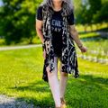A woman with gray hair walking on a gravel path in a sunny park setting, wearing a black t-shirt, patterned cardigan, white pants and brown boots