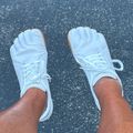 A person wearing white cricket gloves with brown trim stands on a wet asphalt surface, viewed from above showing both feet and legs.