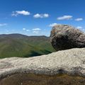 Large boulder balanced on rocky outcrop overlooking forested mountain valley under blue sky with white clouds