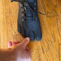 A black toe shoe with individual toe compartments and laces, displayed next to a person's hand for size comparison on a wooden floor