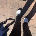 Person's legs and feet walking on brick pavement, wearing dark pants and white shoes, with a bag strap visible and shadows cast on the ground