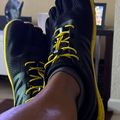 A person wearing black and yellow hiking boots with textured uppers and bright yellow laces, resting their feet up indoors on what appears to be a wooden surface, with a home interior visible in the background.