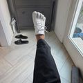 A person wearing black pants and white sneakers sits on a light wood floor in an entryway, with their leg extended toward the camera. Black slippers are visible on the floor nearby, and a dark dresser and white door can be seen in the background.