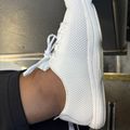 White mesh sneaker with laces, worn on a foot against a gray background.