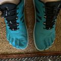 A pair of teal and black athletic running shoes with individual toe compartments, worn by someone standing on a patterned rug