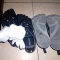 Two pairs of baby booties displayed on a tiled floor. On the left are black booties with white fuzzy trim and white socks. On the right are gray booties with textured soles and black straps.