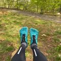Person wearing bright teal toe shoes and black leggings sitting on grass near a wooded path