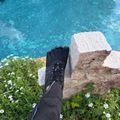 A person wearing a black hiking boot stands on white limestone rocks overlooking turquoise blue water, with white wildflowers and green vegetation visible in the foreground.