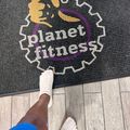 A person wearing white sneakers and blue pants stands on a wooden floor next to a black mat with the Planet Fitness logo, which features a yellow planet design with purple accents and a gear-shaped border.