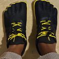 Pair of black and yellow five-finger running shoes with yellow laces, worn by a person standing on a beige floor.