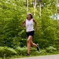 A woman jogging on a rural road surrounded by lush green trees and foliage on a sunny day.
