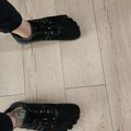A person wearing black athletic shoes with lace-up closures and black pants stands on a light wood-grain laminate floor, shown from above.