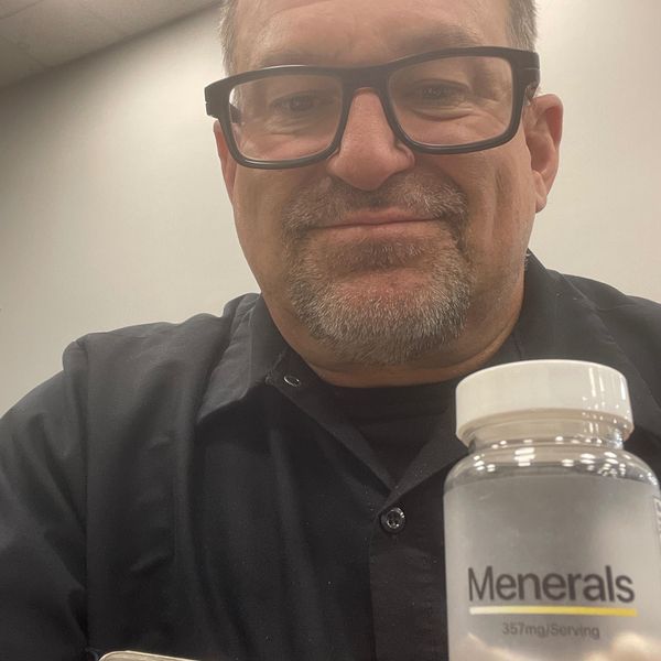 A man wearing glasses and a dark shirt holds a bottle of mineral supplement capsules labeled "Minerals" with "351mg/Serv