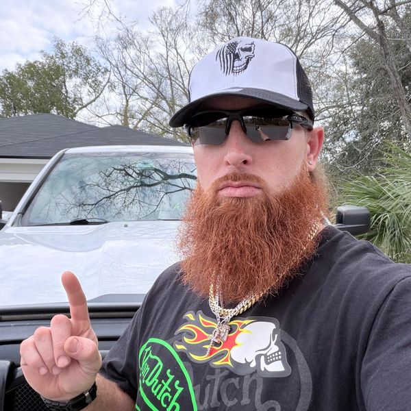 A man with a red beard wearing sunglasses, a baseball cap, and a graphic t-shirt stands in front of a white pickup truck