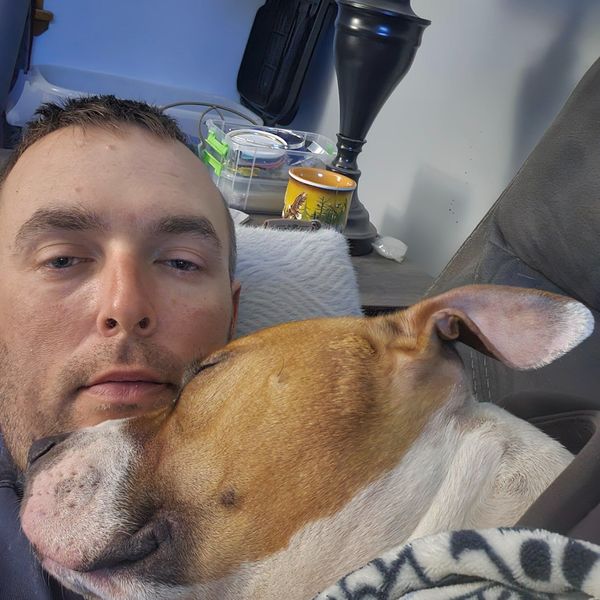 A man taking a selfie while cuddling with a brown and white dog on a couch, with a lamp and yellow mug visible in the ba
