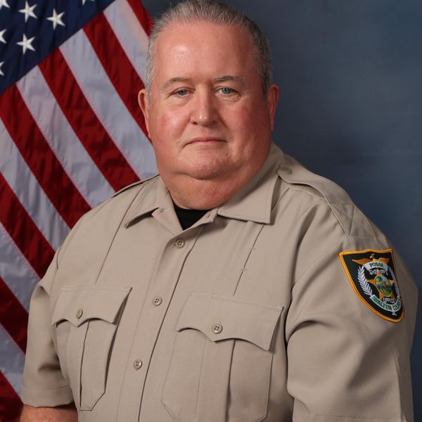 Official portrait of a law enforcement officer in tan uniform standing in front of American flag