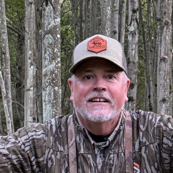 A man in camouflage hunting clothing and cap sitting in a wooded area with trees in the background