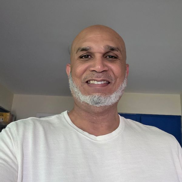 A smiling man with a shaved head and gray beard wearing a white t-shirt, photographed indoors against a gray wall.