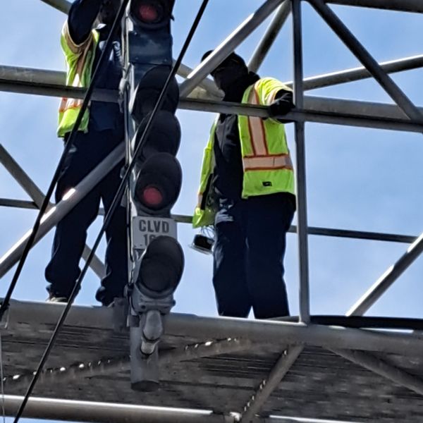 Two workers in high-visibility safety vests performing maintenance on a traffic light mounted on a metal bridge structur