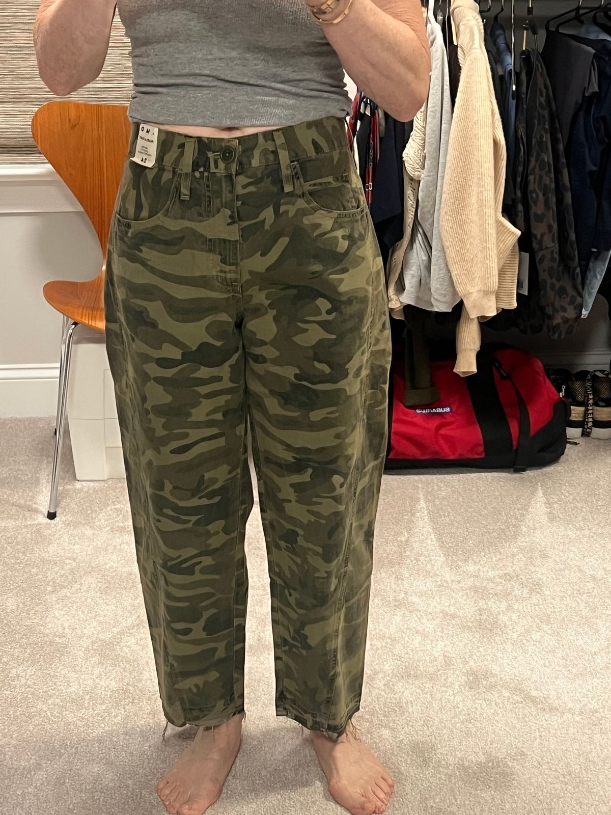 Person wearing a gray tank top and olive green camouflage pants standing in a dressing room with clothing racks visible in the background.