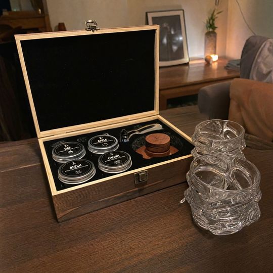 A wooden box containing whiskey smoking wood chips in labeled jars sits next to two crystal glasses on a wooden table