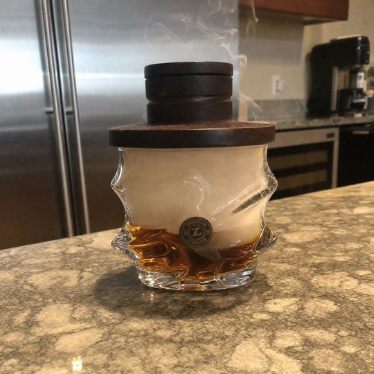 A glass vessel with a wooden lid containing amber liquid sits on a granite countertop in a modern kitchen