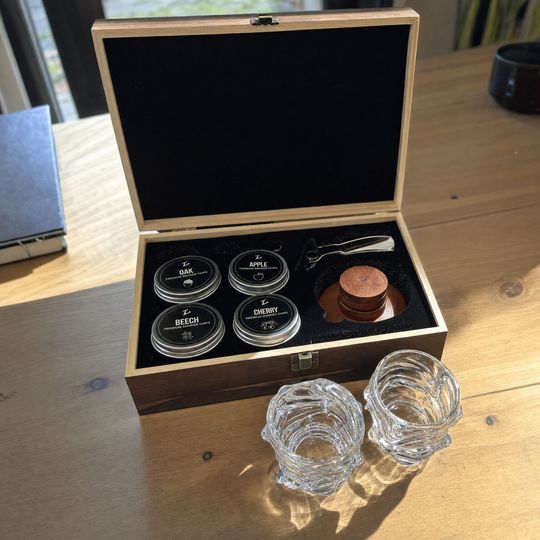A wooden box containing whiskey tasting kit with four labeled containers (Oak, Apple, Beech, Cherry), wooden discs, and a small spoon, displayed alongside two crystal glasses on a wooden table