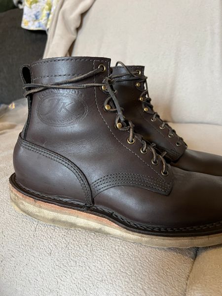 A pair of dark brown leather work boots with laces and light-colored soles sitting on a beige couch