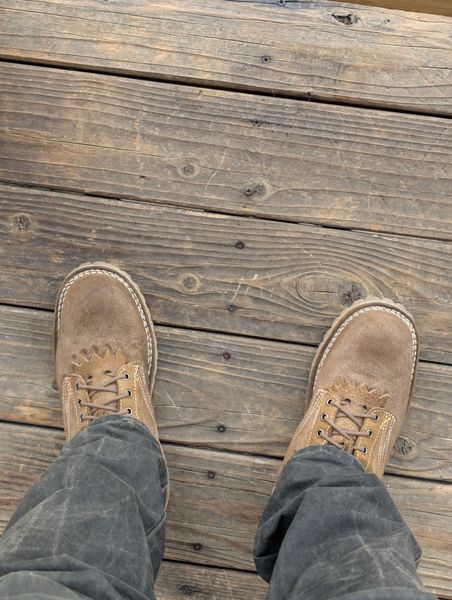 Person wearing brown leather shoes and dark pants standing on weathered wooden deck boards