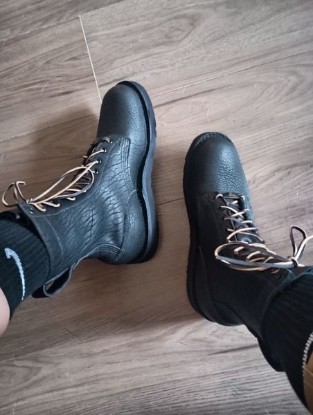 A pair of black leather lace-up shoes on wooden flooring, worn with dark pants