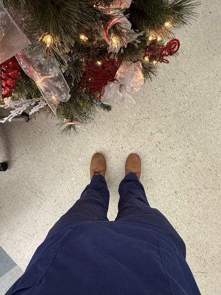 Person wearing navy blue pants and brown shoes standing near a decorated Christmas tree with lights, ribbons, and ornaments on a light colored floor