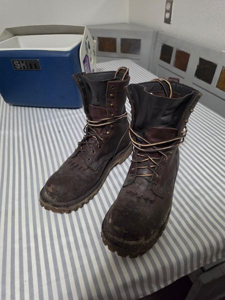 A pair of worn brown leather work boots with laces sitting on a striped bedsheet next to a blue and white cooler
