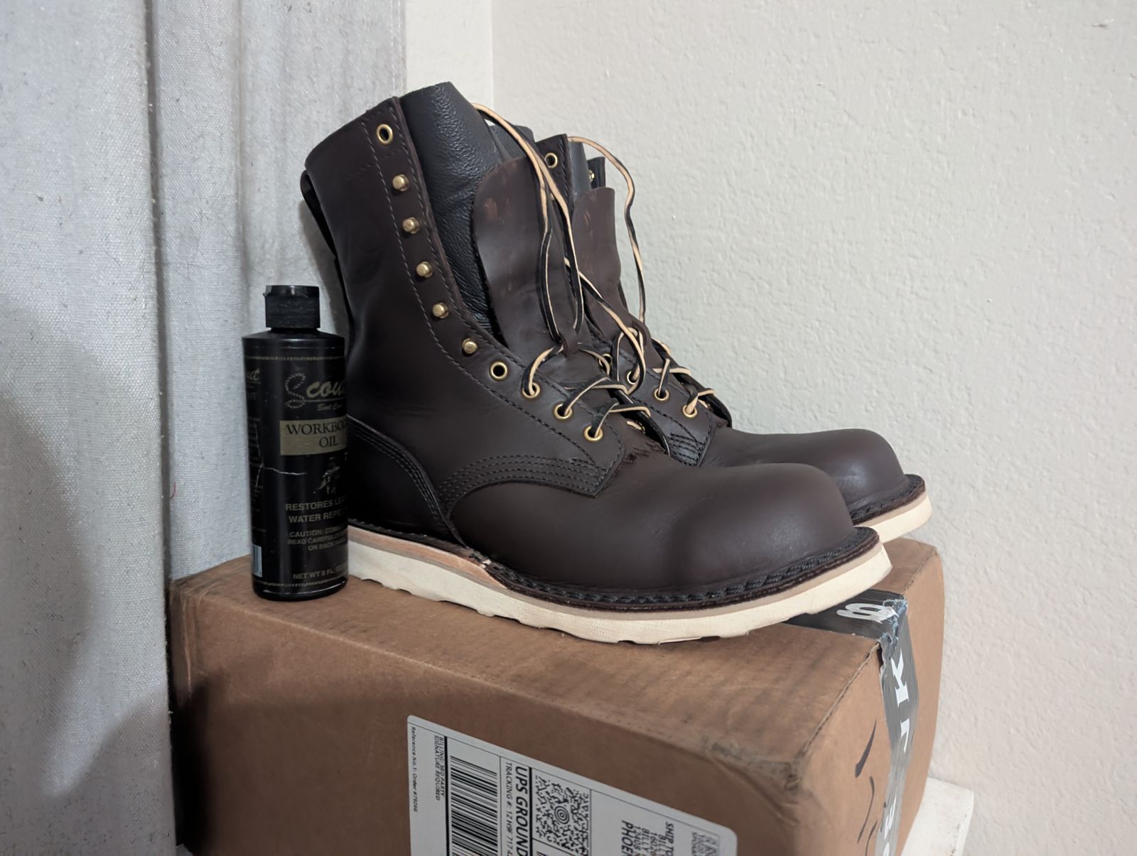Dark brown leather work boots with laces sitting on a cardboard box next to a bottle of leather conditioner