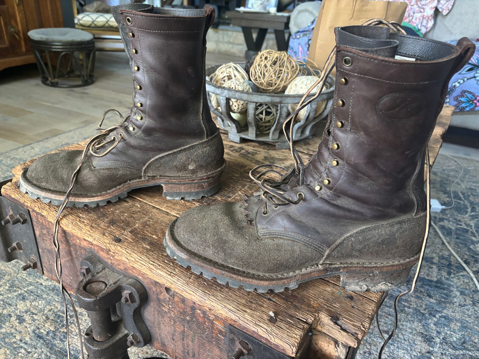 A pair of worn brown leather combat boots with laces sitting on a rustic wooden trunk, with decorative wicker balls visible in a wire basket in the background