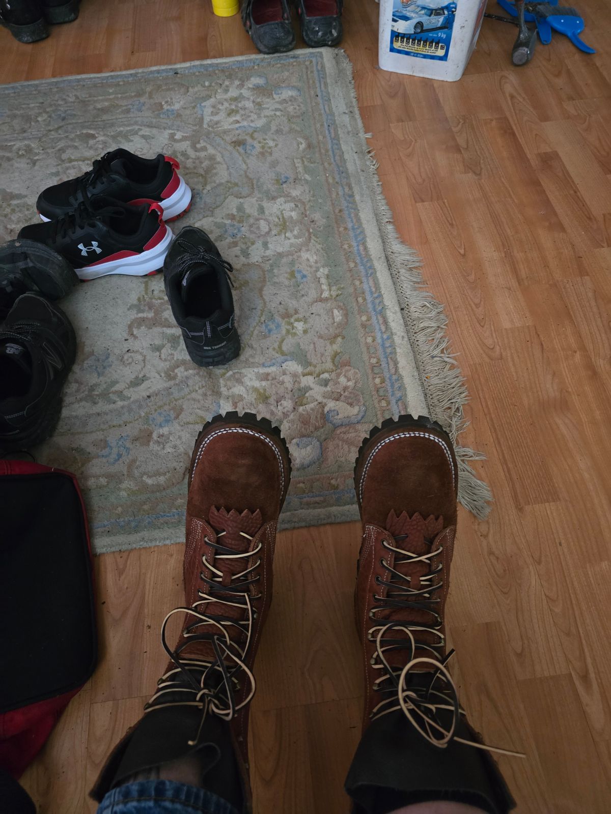 Person wearing brown lace-up boots standing on hardwood floor next to a rug with various shoes scattered around