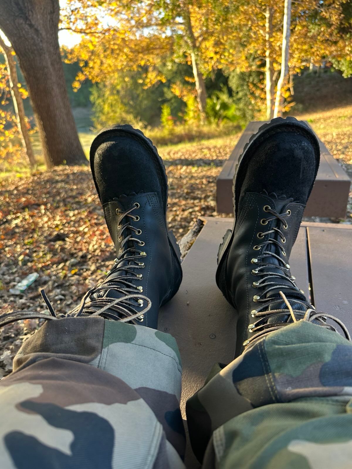 Person wearing black lace-up boots sitting on a bench in an autumn park with golden foliage and fallen leaves