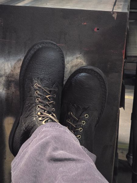 Person wearing black leather lace-up boots with their feet resting on a dark surface