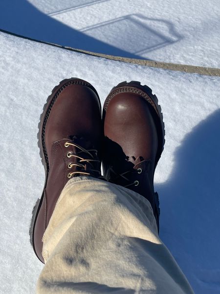 Person wearing brown leather boots with black laces standing on snowy ground with shadows visible
