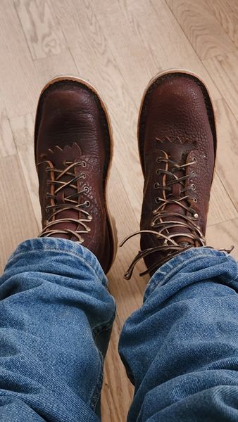 Person wearing brown leather lace-up boots and blue jeans standing on light wooden flooring