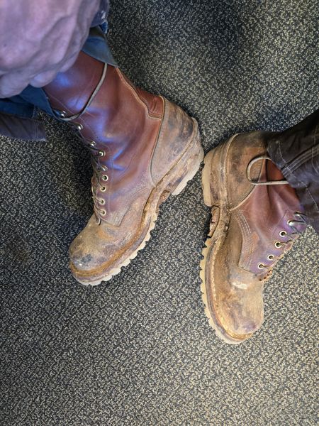 A pair of worn brown leather work boots with laces on a carpeted floor
