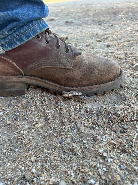 A brown leather boot with laces stepping on sandy ground with small pebbles, worn with blue jeans