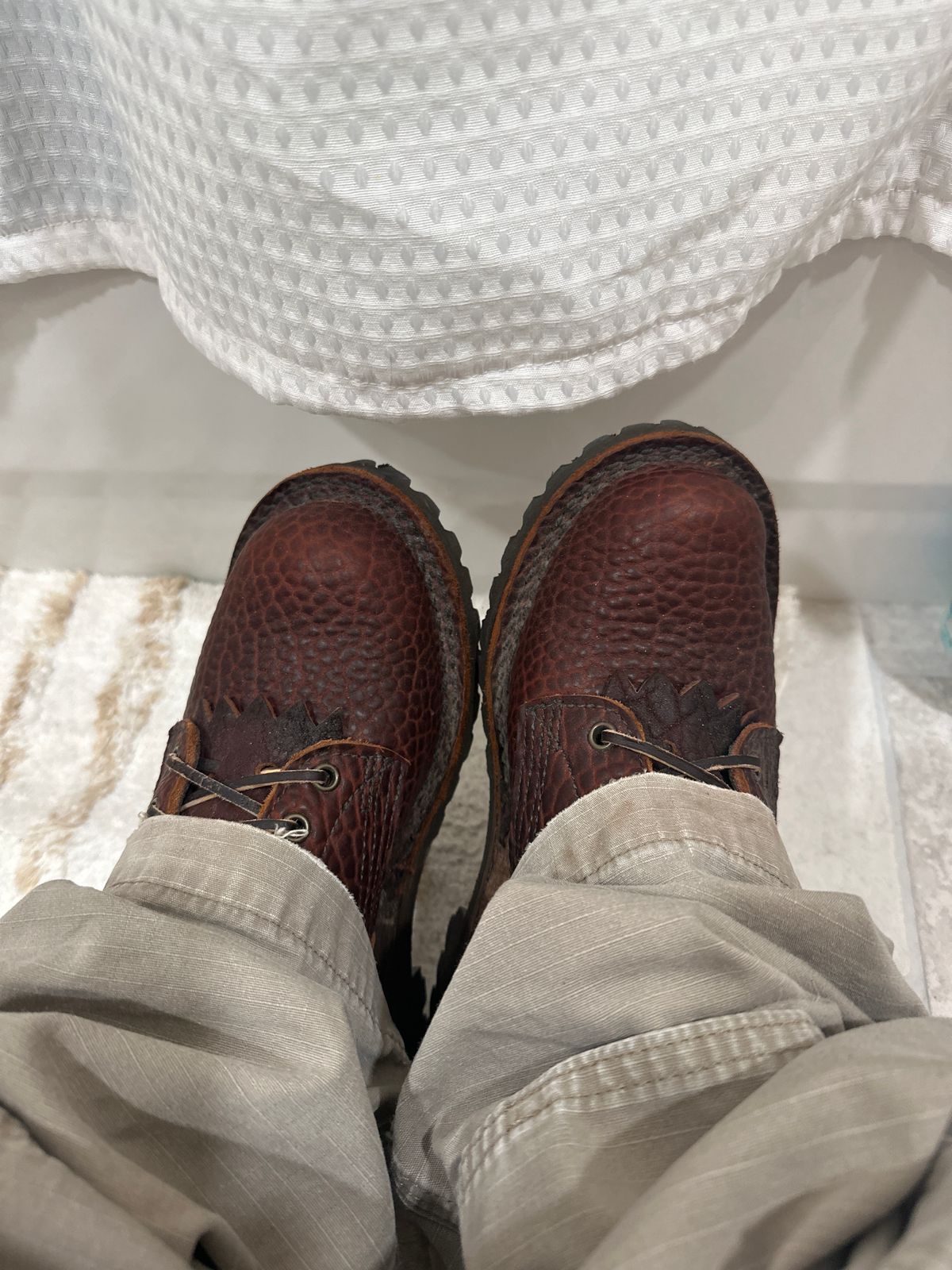 Person wearing brown leather boat shoes and khaki pants sitting with feet up, white textured fabric visible in background
