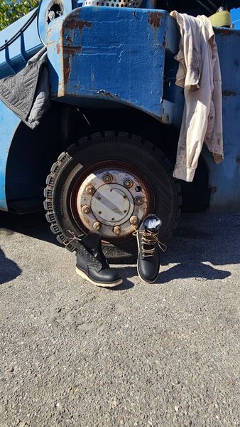 A blue work truck with worn paint parked on asphalt, with work boots and clothing visible near the rear wheel