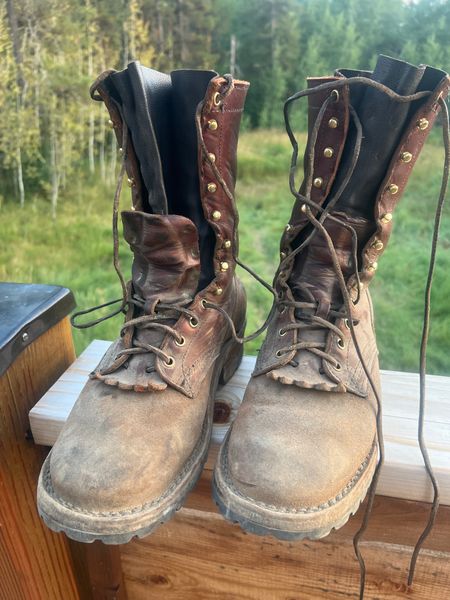 A pair of well-worn brown leather work boots with laces sitting on a wooden deck railing with trees visible in the background