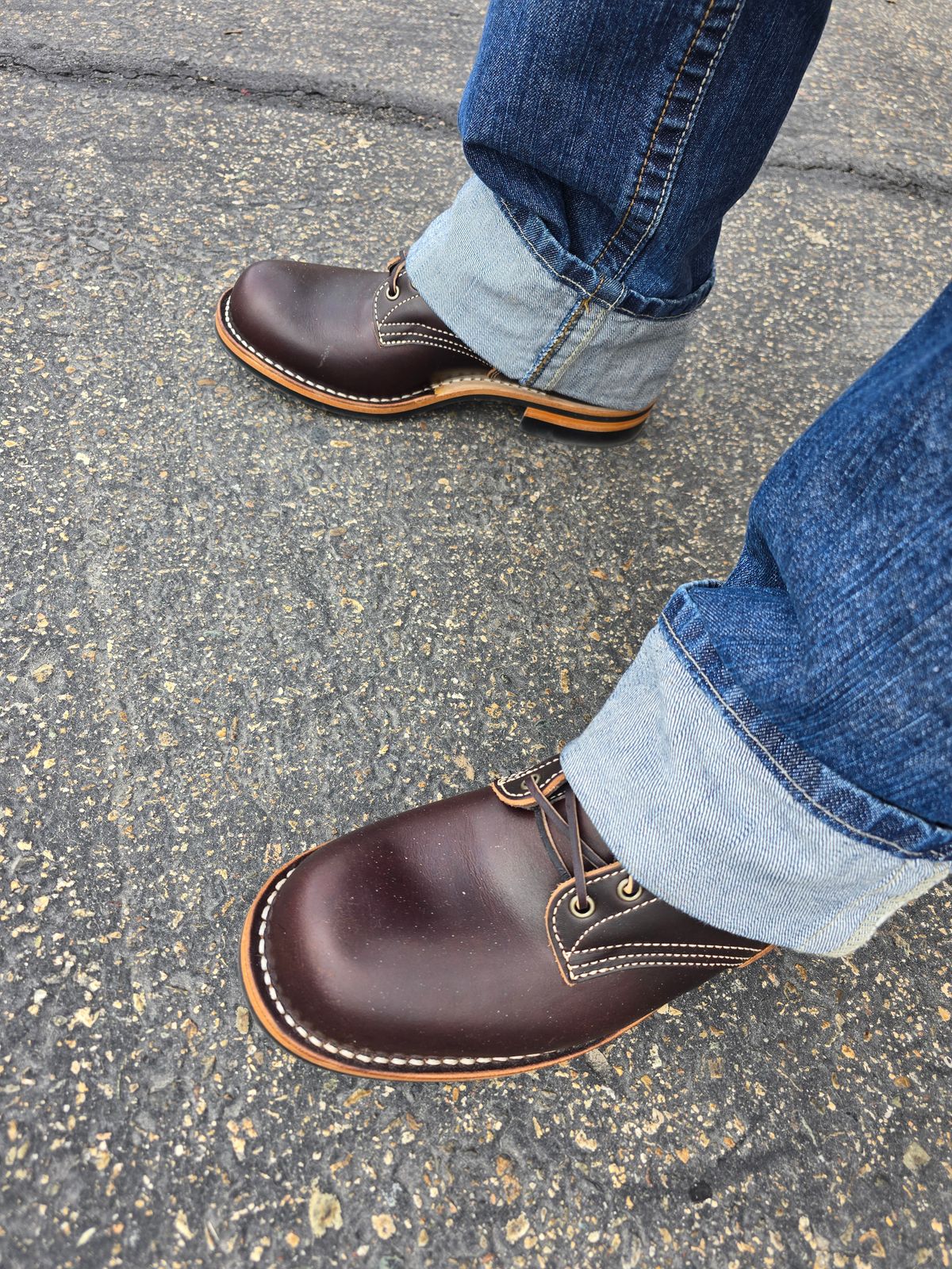 Person wearing dark brown leather shoes with white stitching and cuffed blue jeans, standing on asphalt pavement