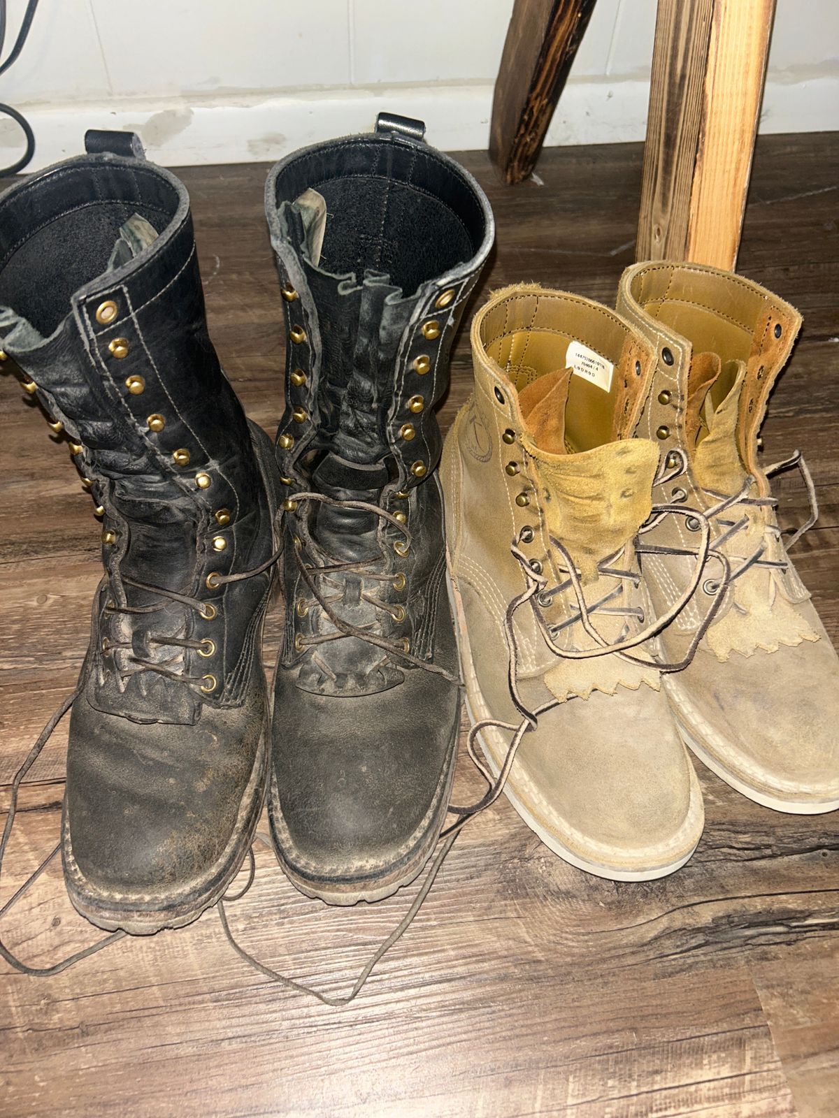 Two pairs of worn work boots on wooden floor - one pair black leather, one pair tan leather, both with laces