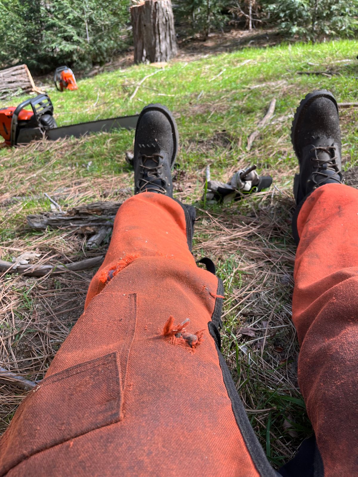 Person wearing orange protective chaps and black boots sitting outdoors with chainsaw equipment visible in background